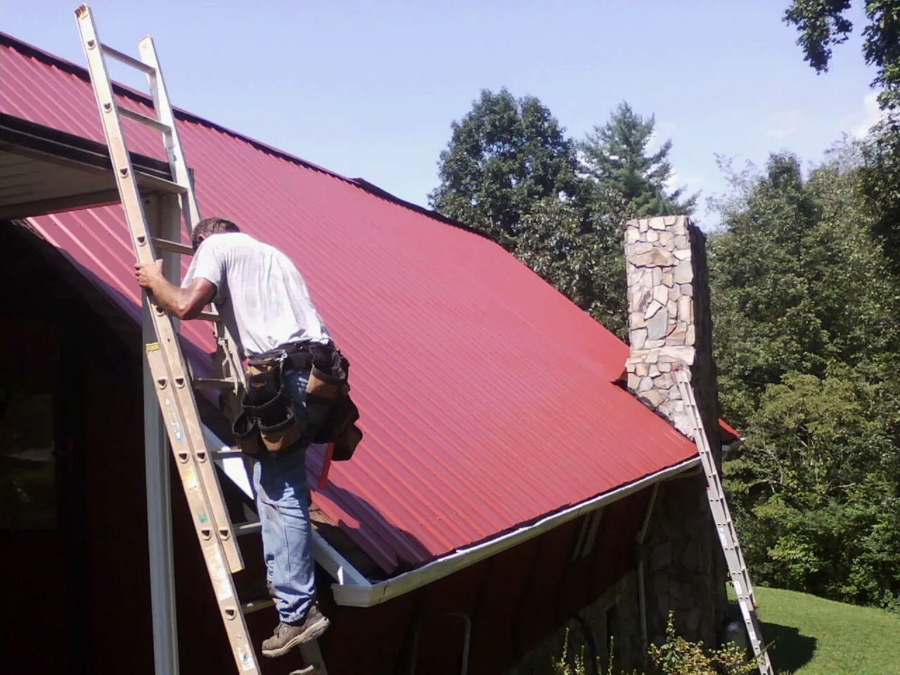 Roofer on a ladder beside a red metal roof and stone chimney — Soffit & Fascia Repair by Roofing Brothers in Barnstead