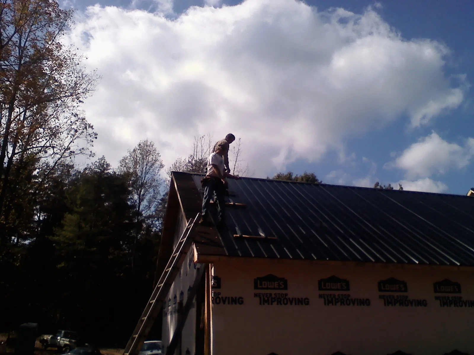 Roofer fastening dark metal panels high against a cloudy sky, from a Roof Inspection job in Barnstead